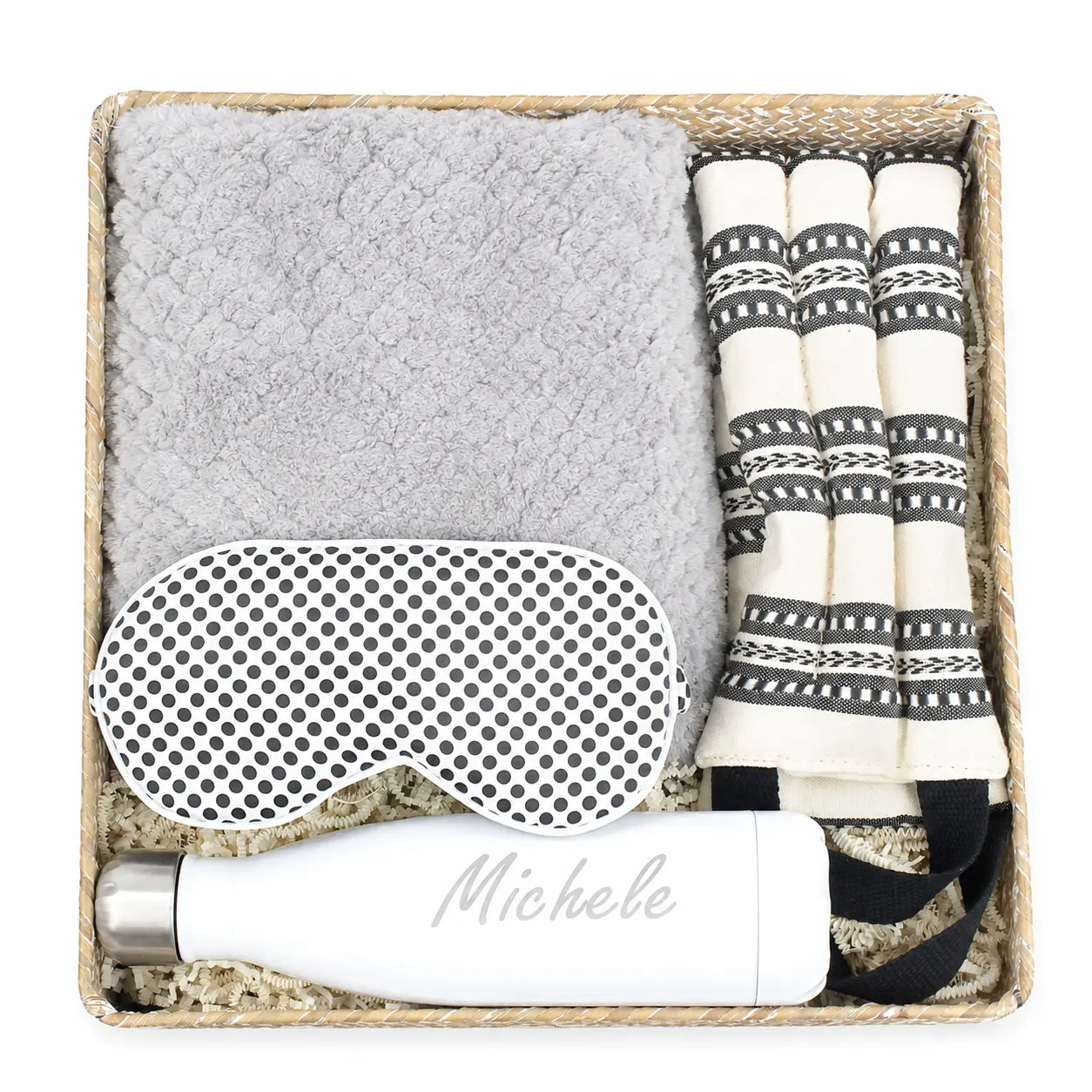 A curated gift set in a woven basket containing a light gray textured blanket, a white sleep mask with black polka dots, a white insulated bottle with the name 'Michele' printed on it, and a set of cream and black patterned fabric accessories.
