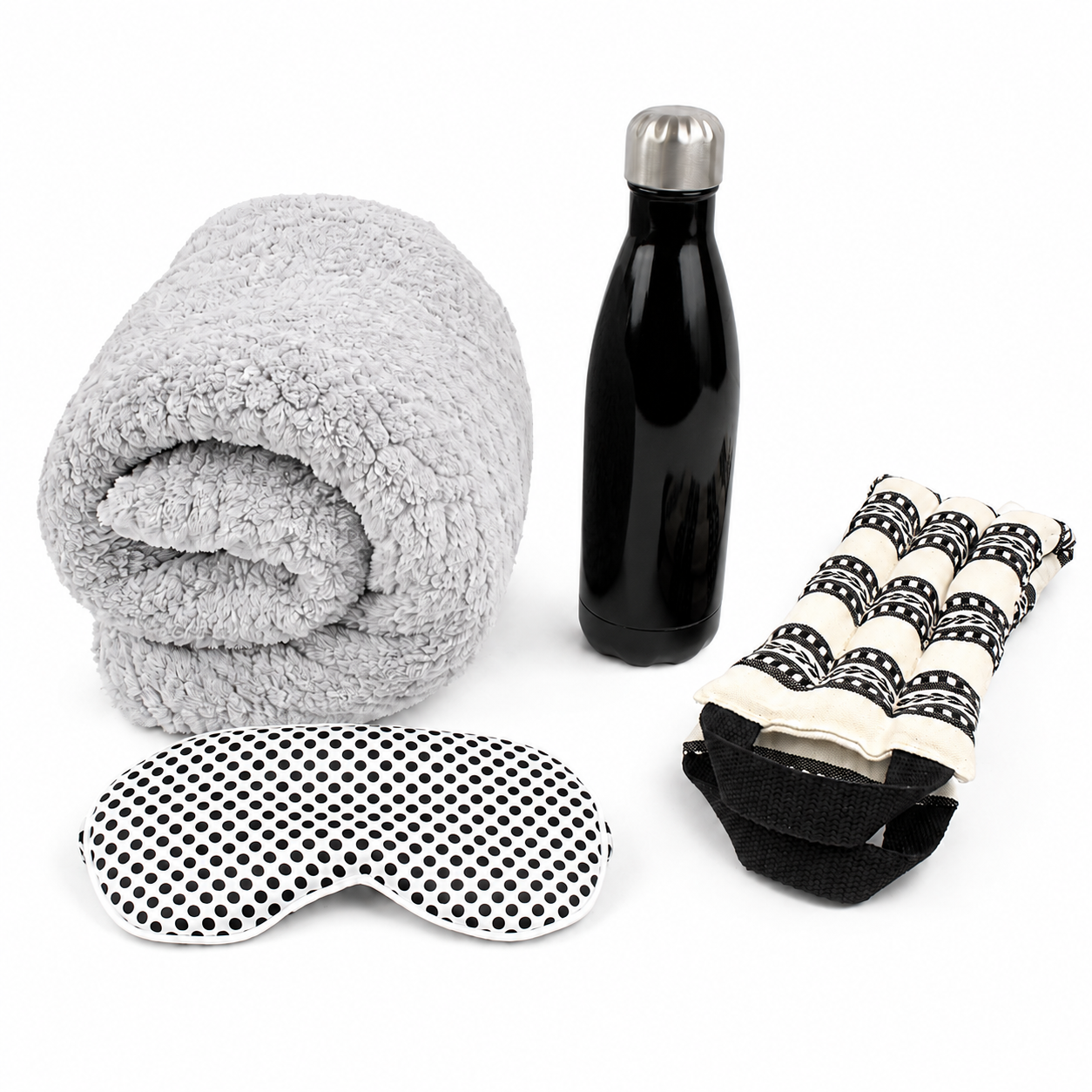 A travel comfort set on a white background, including a rolled light gray sherpa blanket, a black insulated water bottle with a silver cap, a black-and-white polka dot eye mask, and a black-and-white patterned neck pillow.