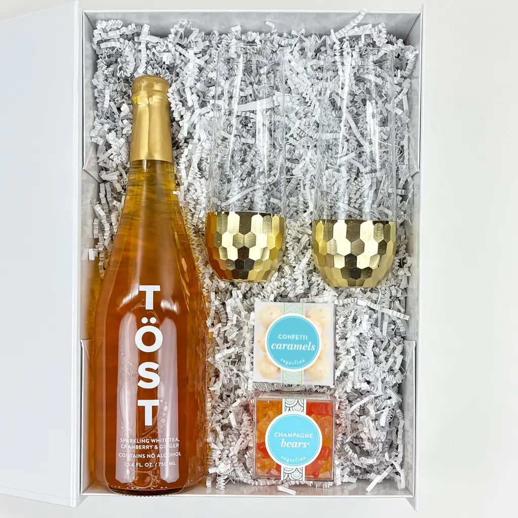 Gift set with a bottle of Töst wine, two gold tumblers, and two boxes of candy in a box with white crinkle paper.