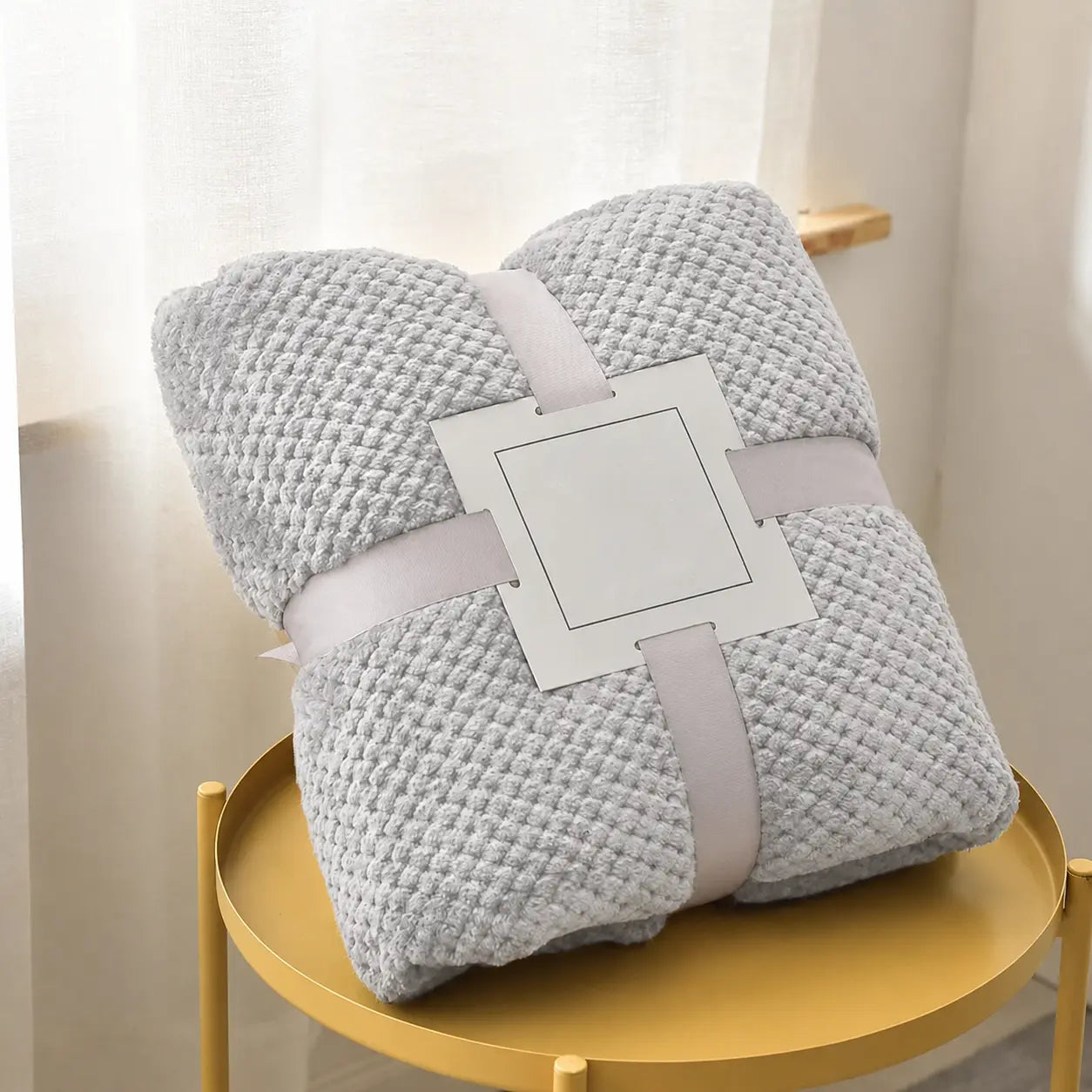 A folded light gray textured throw blanket with a white paper label holder in the center, wrapped in a pale pink ribbon, displayed on a gold metal side table against a soft cream curtain background.