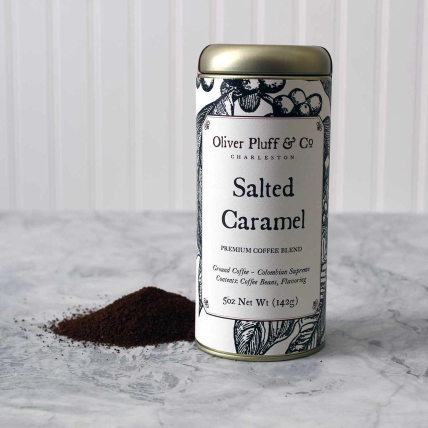 Canister of Oliver Pluff & Co Salted Caramel coffee blend on a marble surface with scattered coffee grounds.