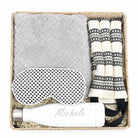 A curated gift set in a woven basket containing a light gray textured blanket, a white sleep mask with black polka dots, a white insulated bottle with the name 'Michele' printed on it, and a set of cream and black patterned fabric accessories.