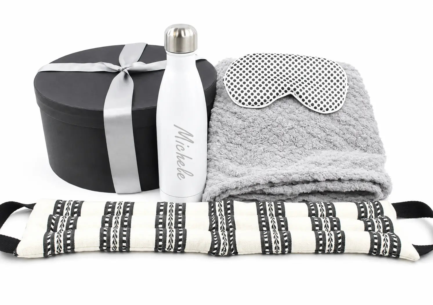 A curated gift set on a white background featuring a black round presentation box with a silver ribbon, a white insulated water bottle personalized with 'Michelle' in silver script, a folded gray plush towel, a gray and white polka-dot eye mask, and a black-and-white patterned neck pillow.