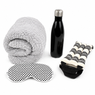 A travel comfort set on a white background, including a rolled light gray sherpa blanket, a black insulated water bottle with a silver cap, a black-and-white polka dot eye mask, and a black-and-white patterned neck pillow.