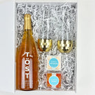 Gift set with a bottle of Töst wine, two gold tumblers, and two boxes of candy in a box with white crinkle paper.