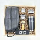 Grooming kit with leather bag, shaving brush, face scrub, pomade, and tumbler in a wooden box.