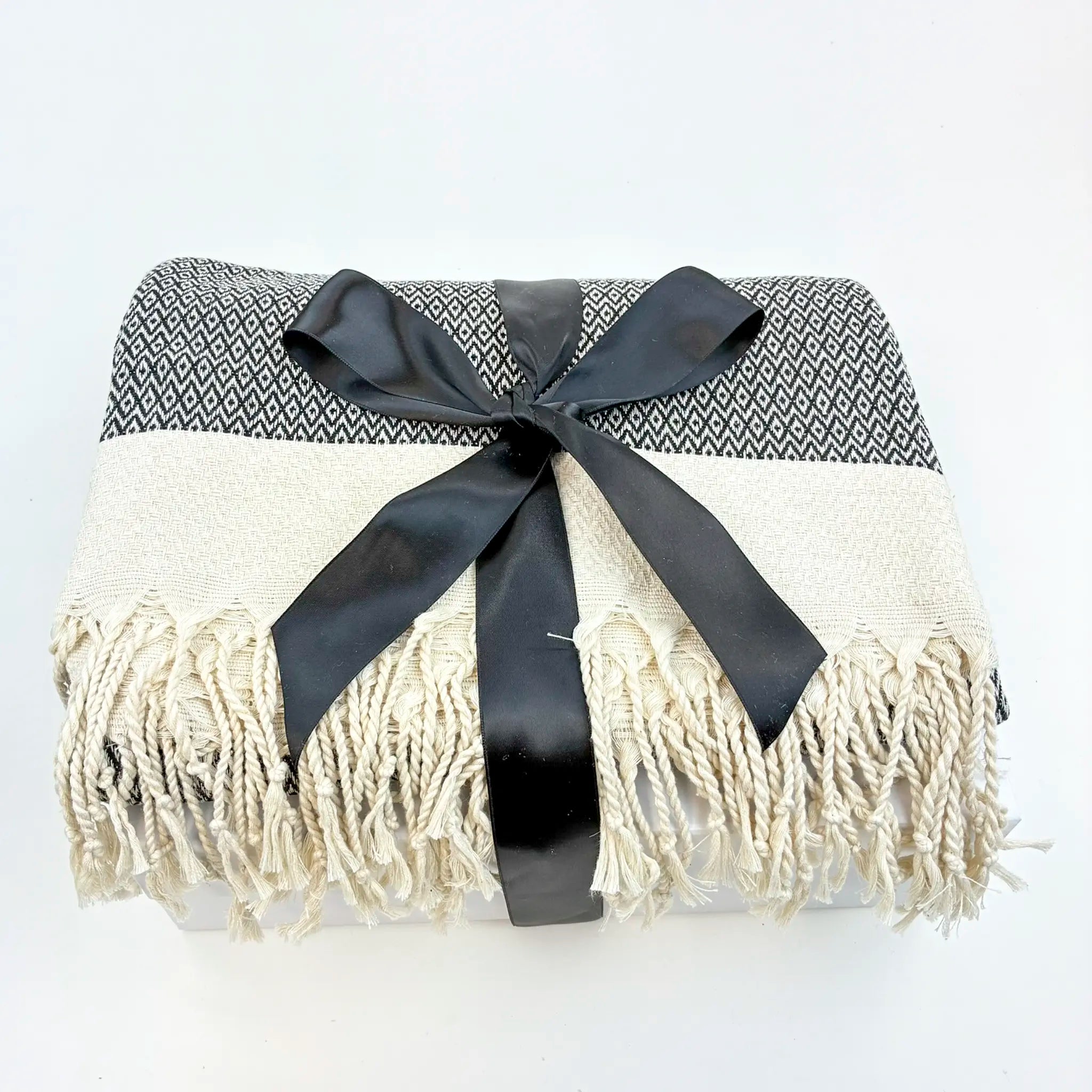 Blanket with black and white pattern and white fringe, tied with a black ribbon on a light gray background.