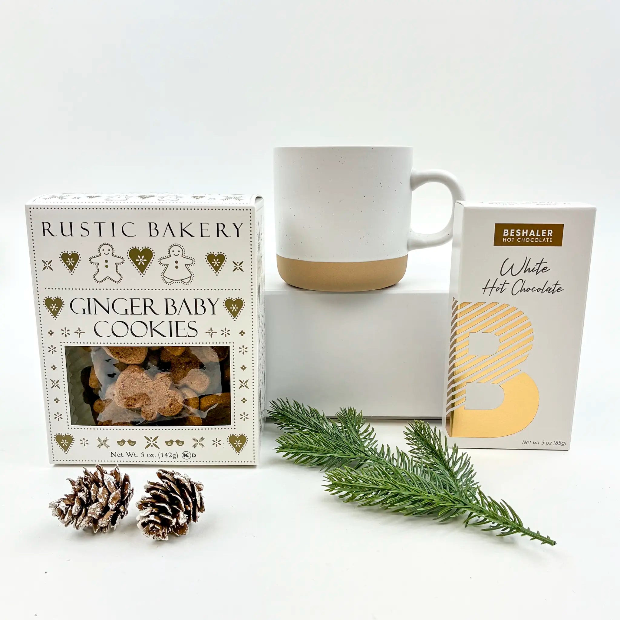 Box of 'Ginger Baby Cookies' from Rustic Bakery, a mug, and a package of Beshaler White Hot Chocolate on a white background.